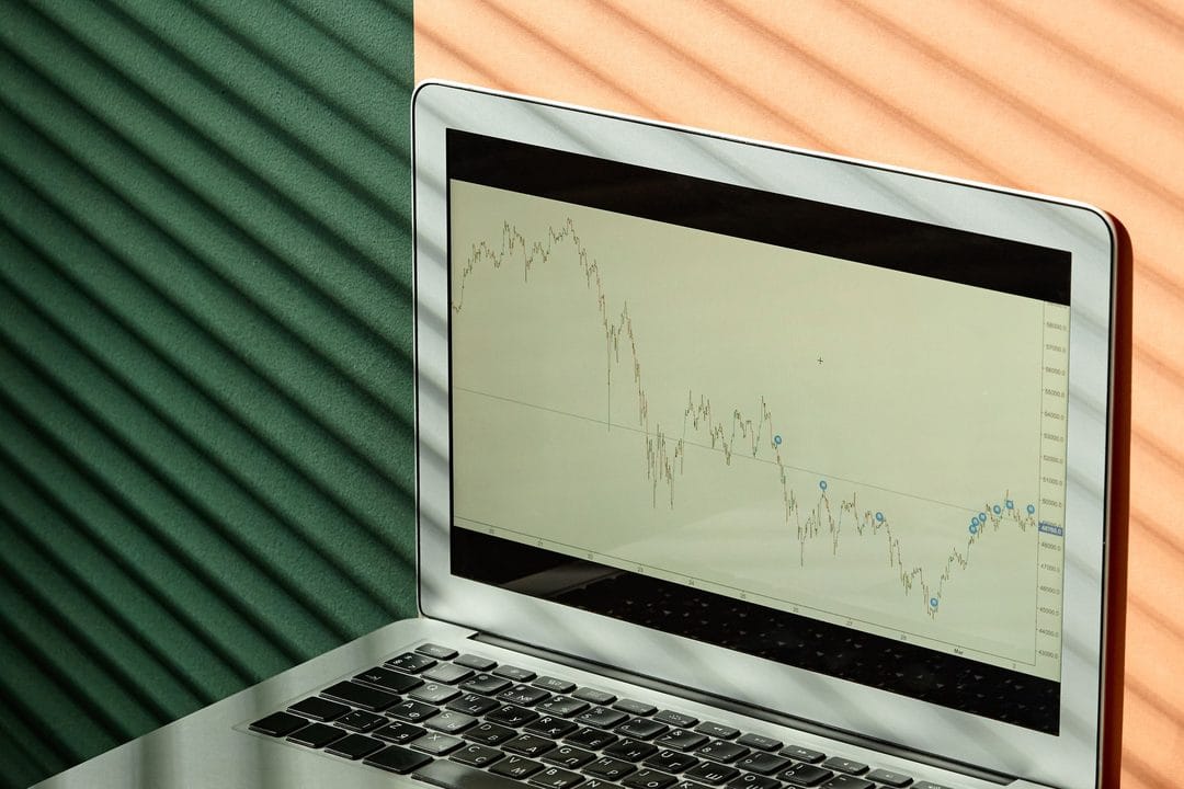 Laptop screen showing a stock market chart alongside printed finance reports, used to illustrate reviewing an investment portfolio